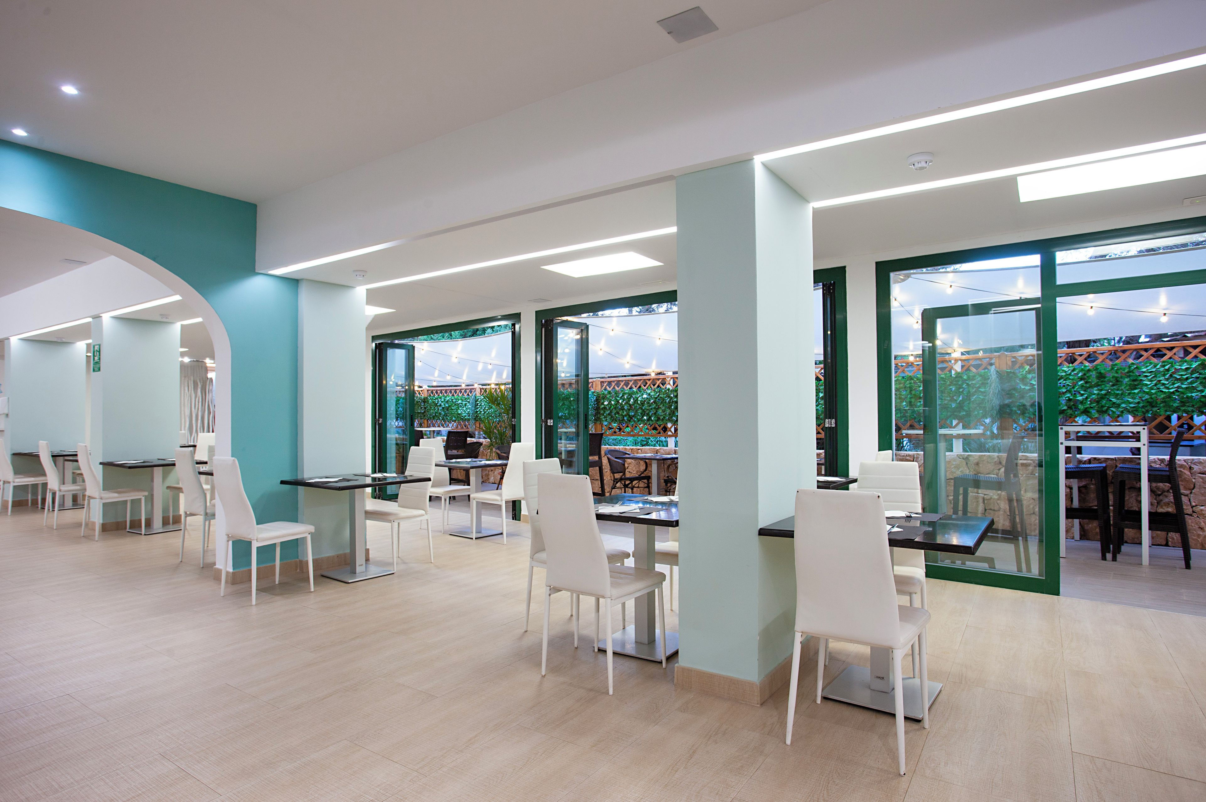 A modern dining area with white chairs and tables, large windows with green frames, and a view of an outdoor area with string lights.