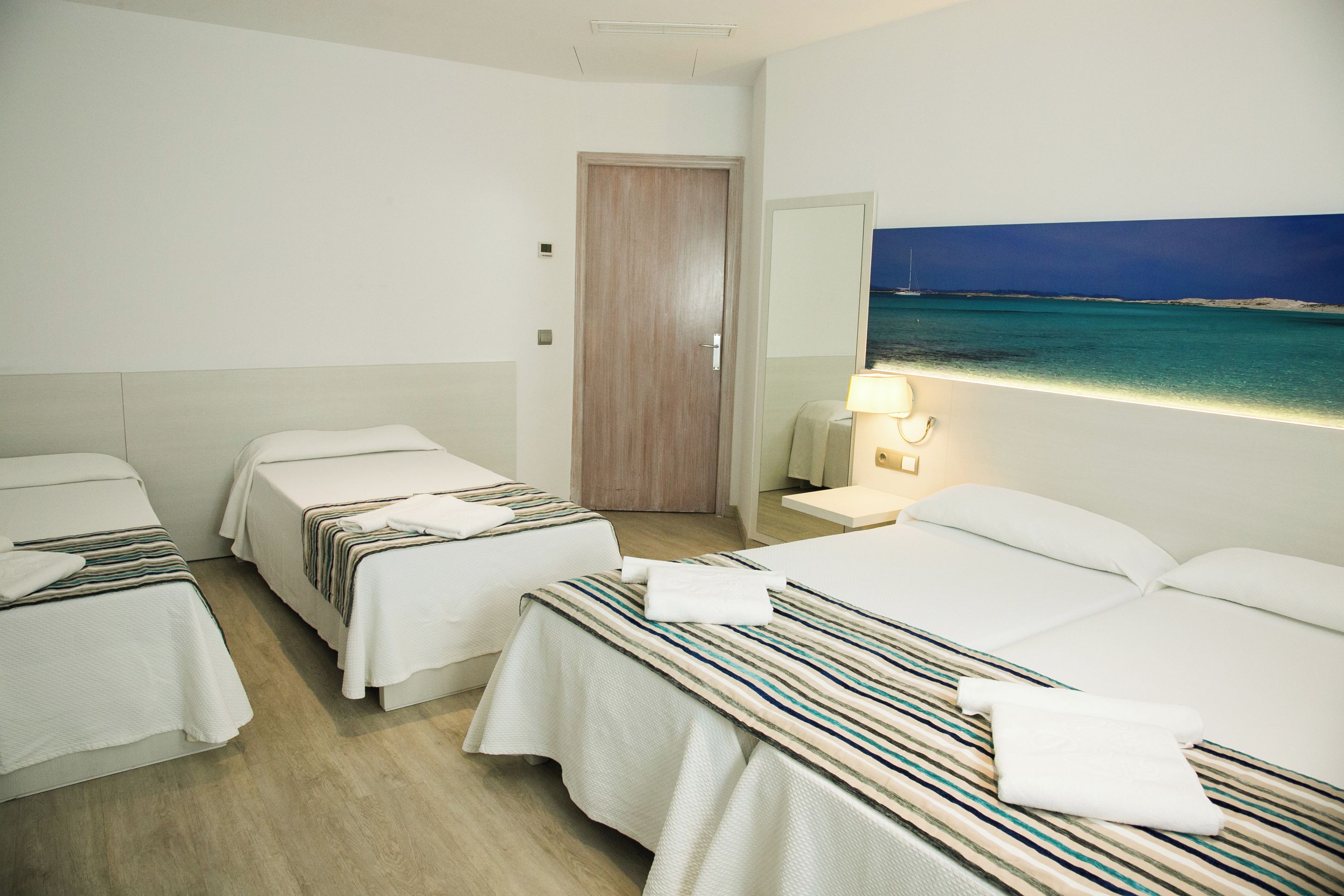 Habitación de hotel con tres camas, una puerta de madera, un cuadro de una playa y una lámpara de noche.