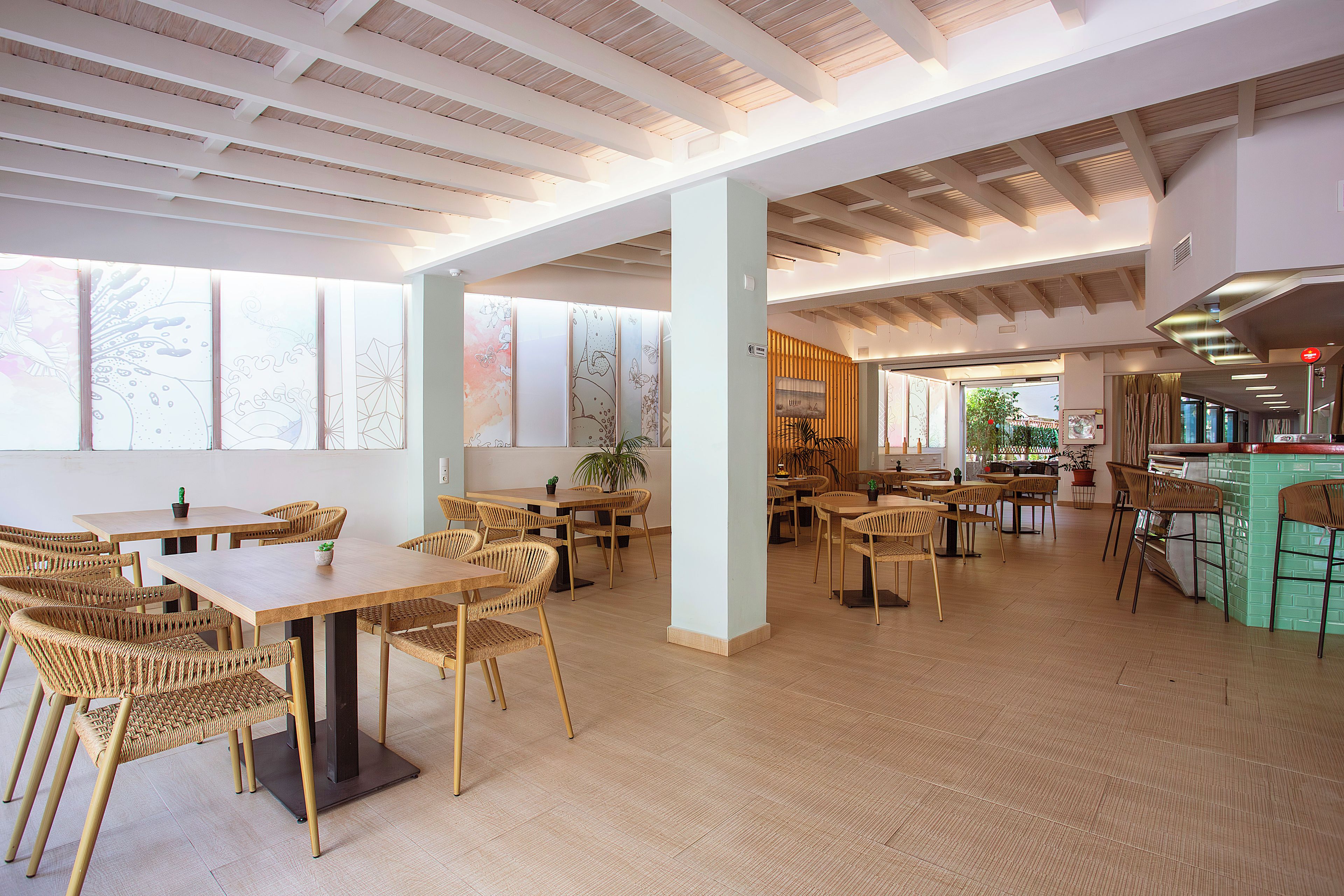 Un espacio interior amplio con varias mesas y sillas de madera, ventanas grandes y una zona de cocina moderna.