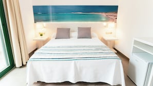 Ein Hotelzimmer mit einem Bett, zwei Nachttischlampen, einem Nachttisch, einem Fenster mit Vorhängen und einem an der Wand montierten Fernseher.