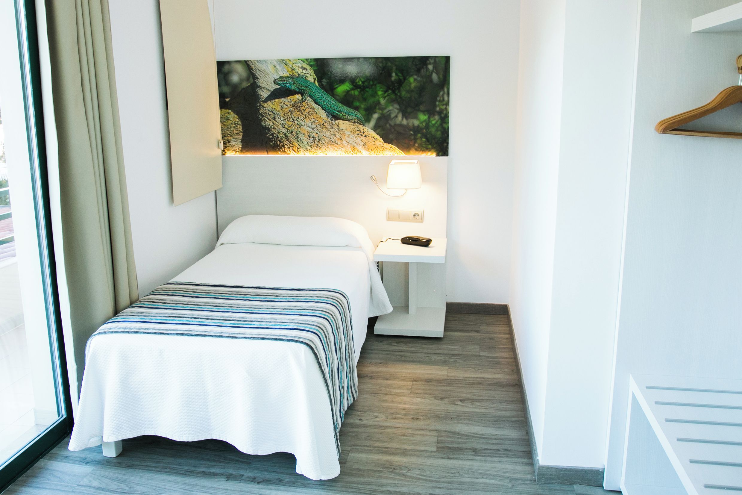 A hotel room with a bed, a nightstand, a lamp, a painting of a lizard, and a wooden hanger.