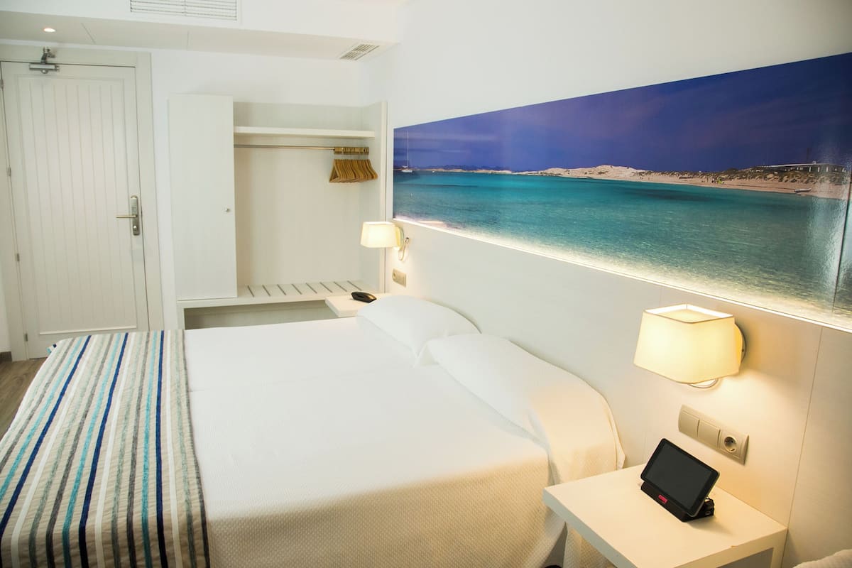 Une chambre d’hôtel avec un lit, une table de chevet avec une lampe, un tableau représentant une plage et une armoire.