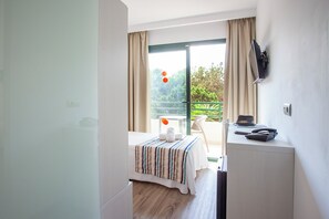 Egy szállodai szoba, amelyben található egy ágy, egy íróasztal, egy szék és egy balkon, ahonnan zöld területre nyílik kilátás.