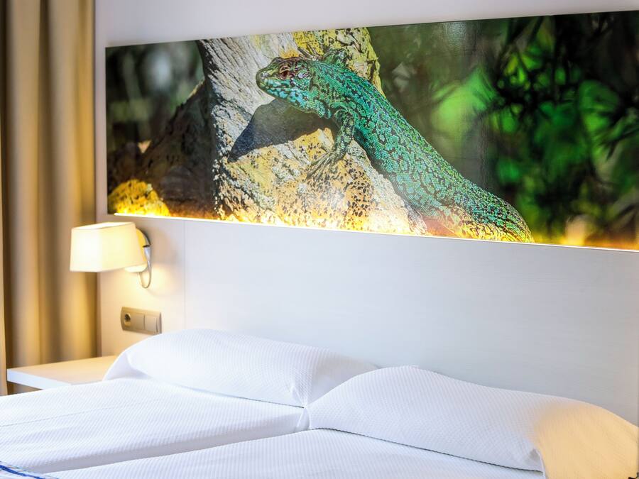 Una habitación de hotel con una cama, una lámpara de pared y un cuadro enmarcado de un lagarto en la pared.