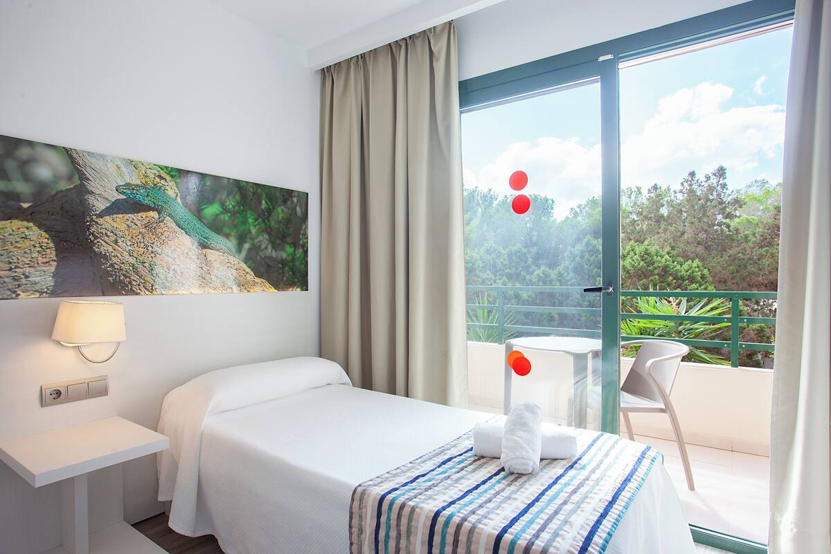 Une chambre d’hôtel avec un lit, une table de chevet, une lampe, une fenêtre avec des rideaux et une vue sur la verdure.