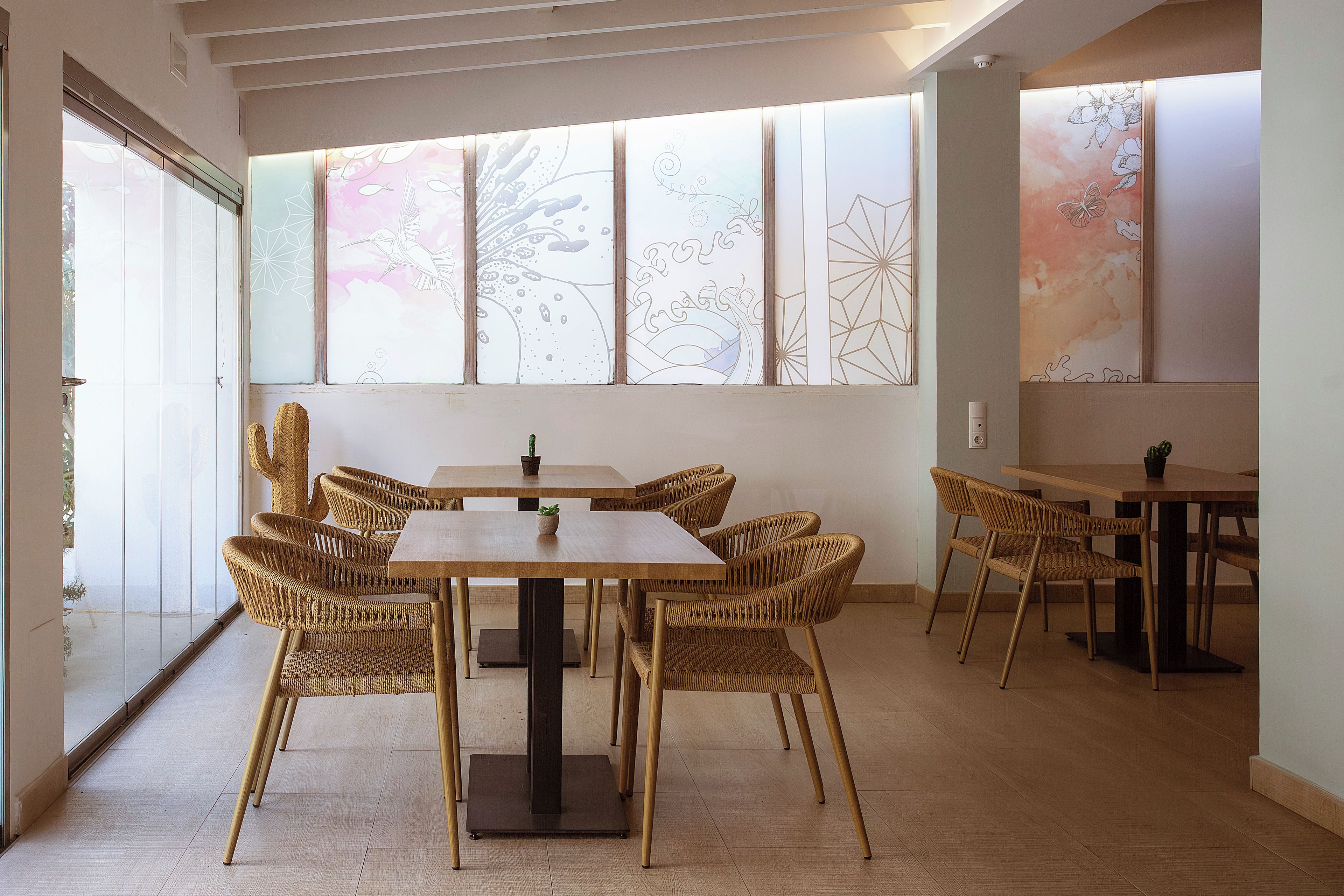 A dining area with a wooden table and wicker chairs, large windows with decorative patterns, and a light-colored floor.