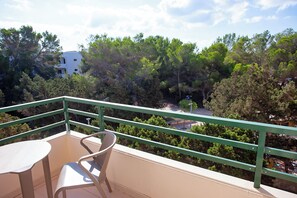A balcony with a table and chair, overlooking a densely wooded area and a building in the background. - Hotel Rosales Formentera (Formentera)