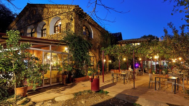 An outdoor cafe with tables and chairs, a building with arched windows, and potted plants. - Mas Torrencito (Vilademuls)
