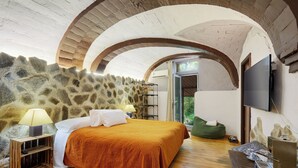 A bedroom with a stone wall, wooden floor, a bed with an orange bedspread, a nightstand with a lamp, a green bean bag chair, and a TV mounted on the wall. - Mas Torrencito (Vilademuls)