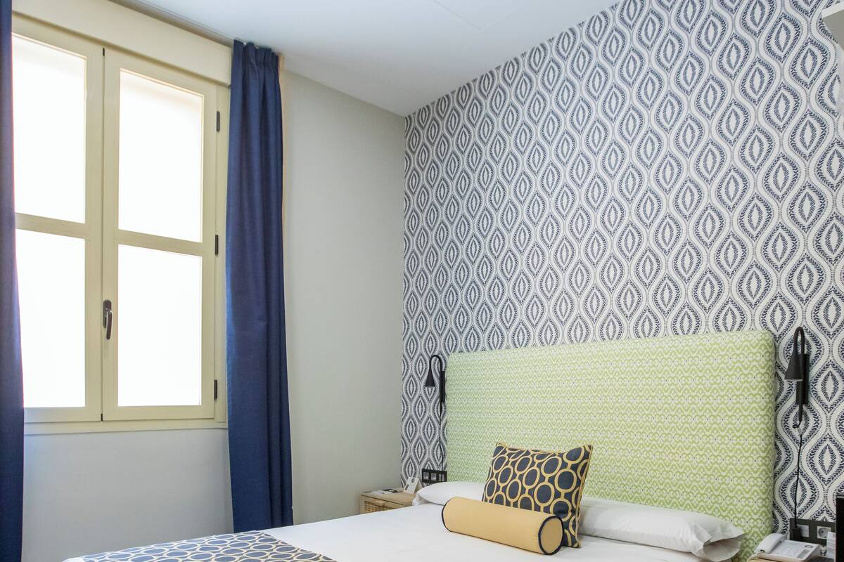 Ein ordentlich bezogenes Bett mit gemusterten Bettwäsche, ein Nachttisch mit Lampe und eine Wand mit Muster.