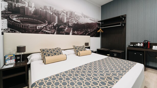 Ein modernes Hotelzimmer mit einem großen Bett, einem Wandgemälde mit Stadtansicht, einem Schreibtisch mit Kaffeemaschine und einem an der Wand befestigten Kleiderhaken.