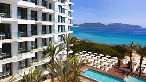 Ett modernt hotell med flera balkonger, ett poolområde med solstolar och utsikt över havet.