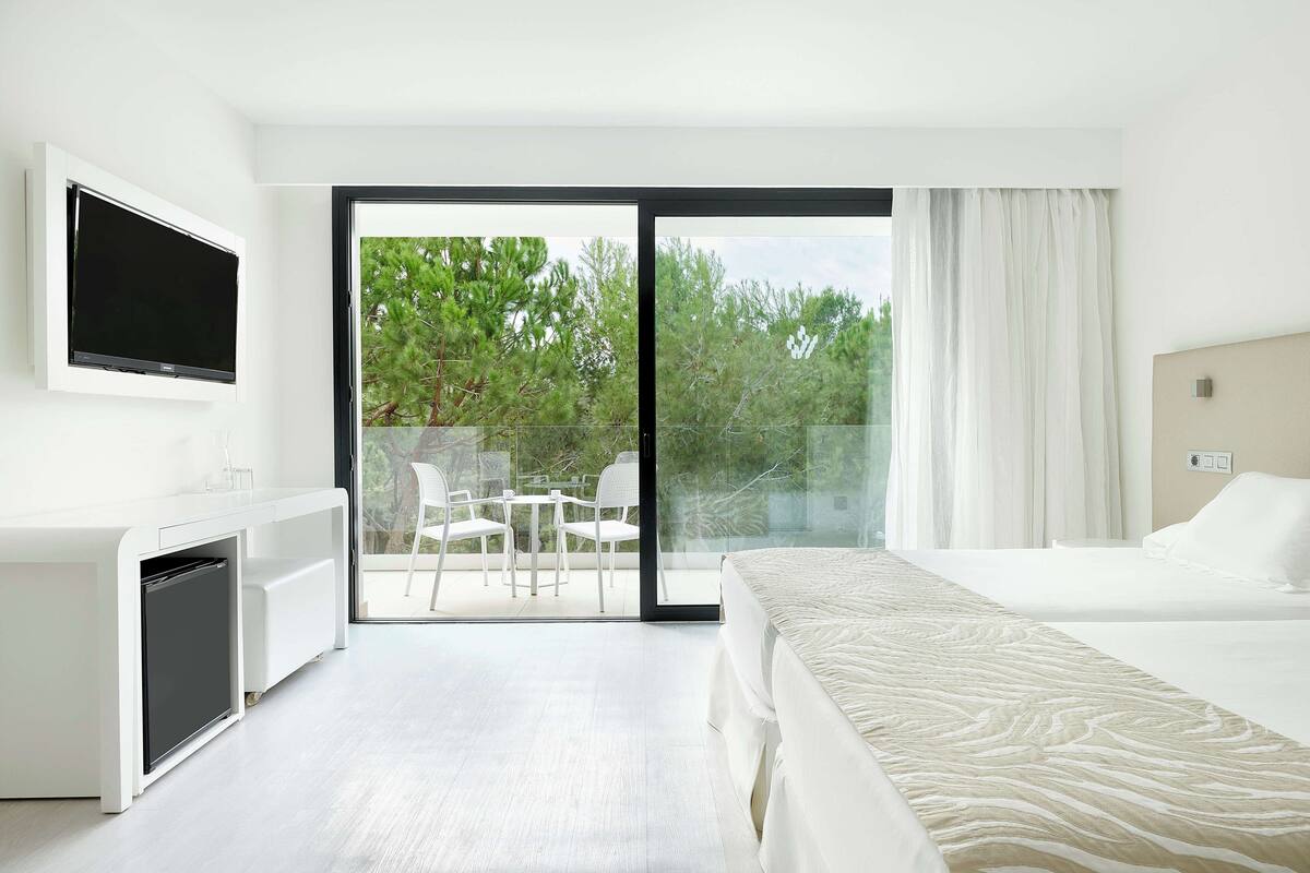 Quarto de hotel com uma cama, uma escrivaninha com televisão, uma porta de correr para uma varanda com cadeiras e vista para árvores.
