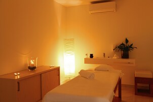 Un dormitorio con una cama, una mesita de noche con una vela, una mesita auxiliar y una planta en maceta, además de un aire acondicionado instalado en la pared.