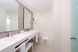 Un baño moderno con un espejo grande, dos lavamanos, una ducha y un pequeño banco blanco.