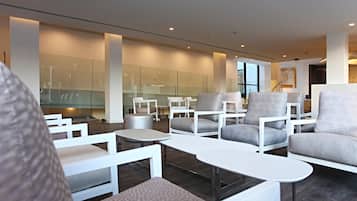 A modern lounge area with white furniture, glass partitions, and a staircase in the background.