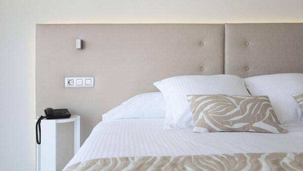 A neatly made bed with a patterned throw, a bedside phone, and a wall-mounted light switch. - Iberostar Waves Cala Millor (Sant Llorenc des Cardassar)