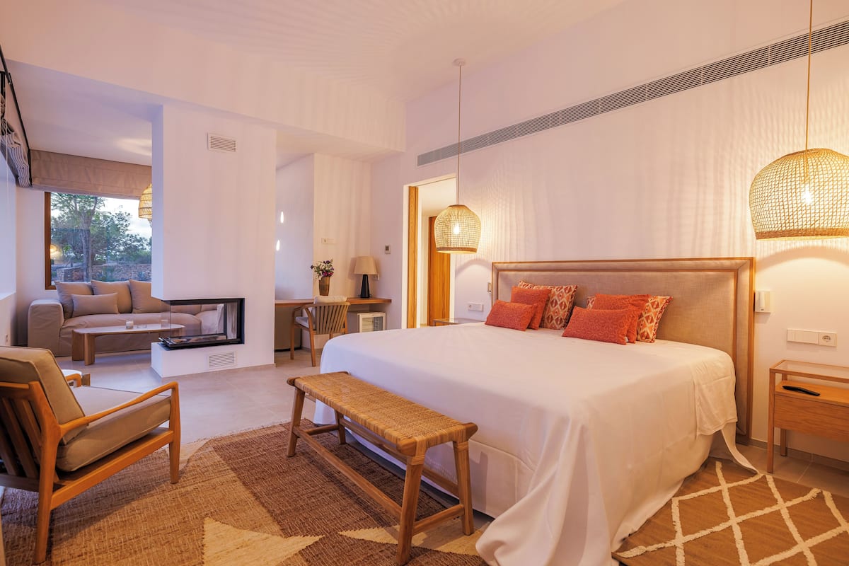 A modern hotel room with a large bed, a sitting area with a sofa and armchair, a small desk, and a large window.