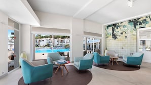 A spacious lounge area with teal armchairs, a brown rug, and a large window overlooking a pool and buildings. - TRH Tirant Playa (Mercadal)