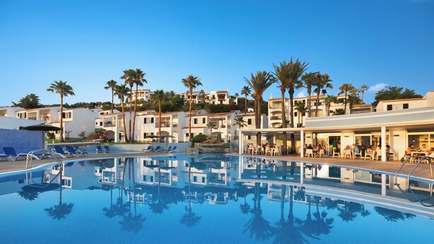A swimming pool with clear blue water, surrounded by lounge chairs and palm trees. - TRH Tirant Playa (Mercadal)
