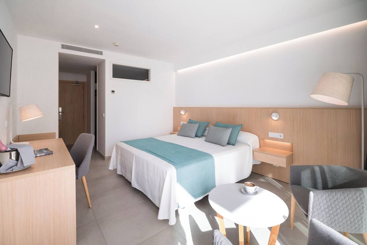 Una habitación de hotel moderna con una cama bien hecha, un escritorio con lámpara, una silla y una mesita con una taza.