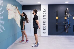 Dos personas interactuando con un mapa colgado en la pared de una habitación, con un letrero 'BIKE FRIENDLY', una bicicleta montada en la pared y una puerta al fondo.