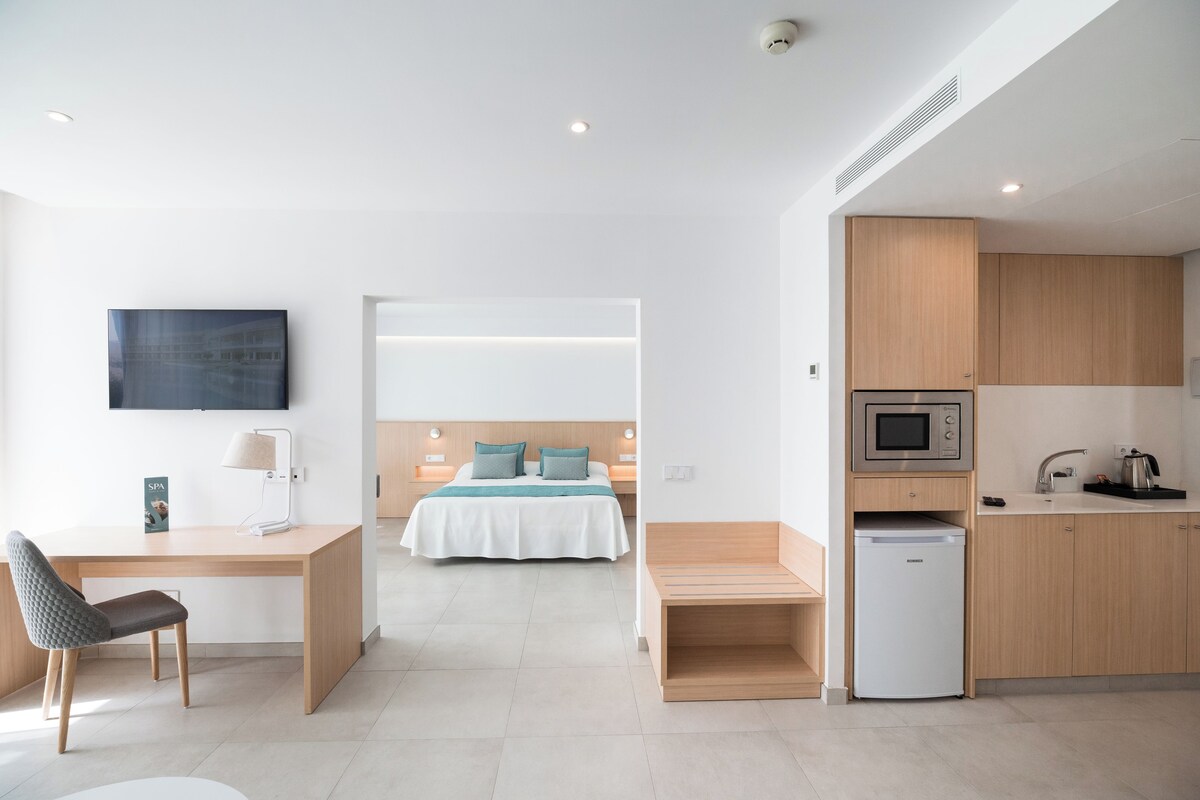 A modern hotel room with a bed, desk, chair, and kitchenette.