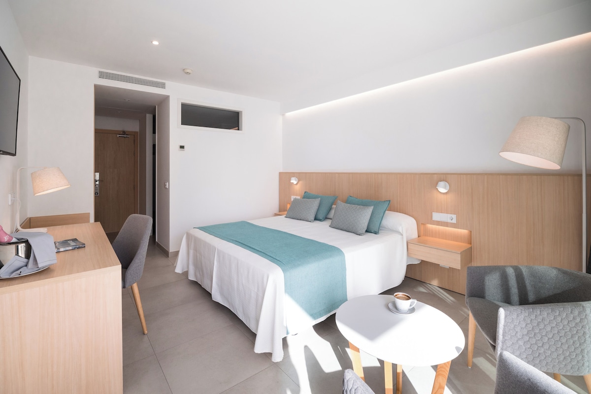 Una habitación de hotel moderna con una cama bien hecha, un escritorio con lámpara, una silla y una mesita con una taza.