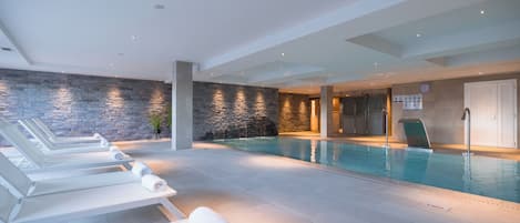 A modern indoor swimming pool with lounge chairs and a stone wall.