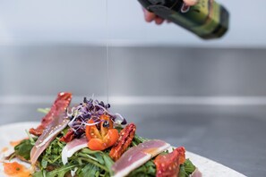 Primer plano de un plato con diversos ingredientes, entre ellos verduras, tomates y carne, que se está preparando en una cocina.