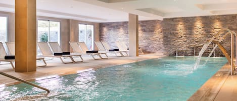 An indoor swimming pool with clear water, sun loungers, and a stone wall.