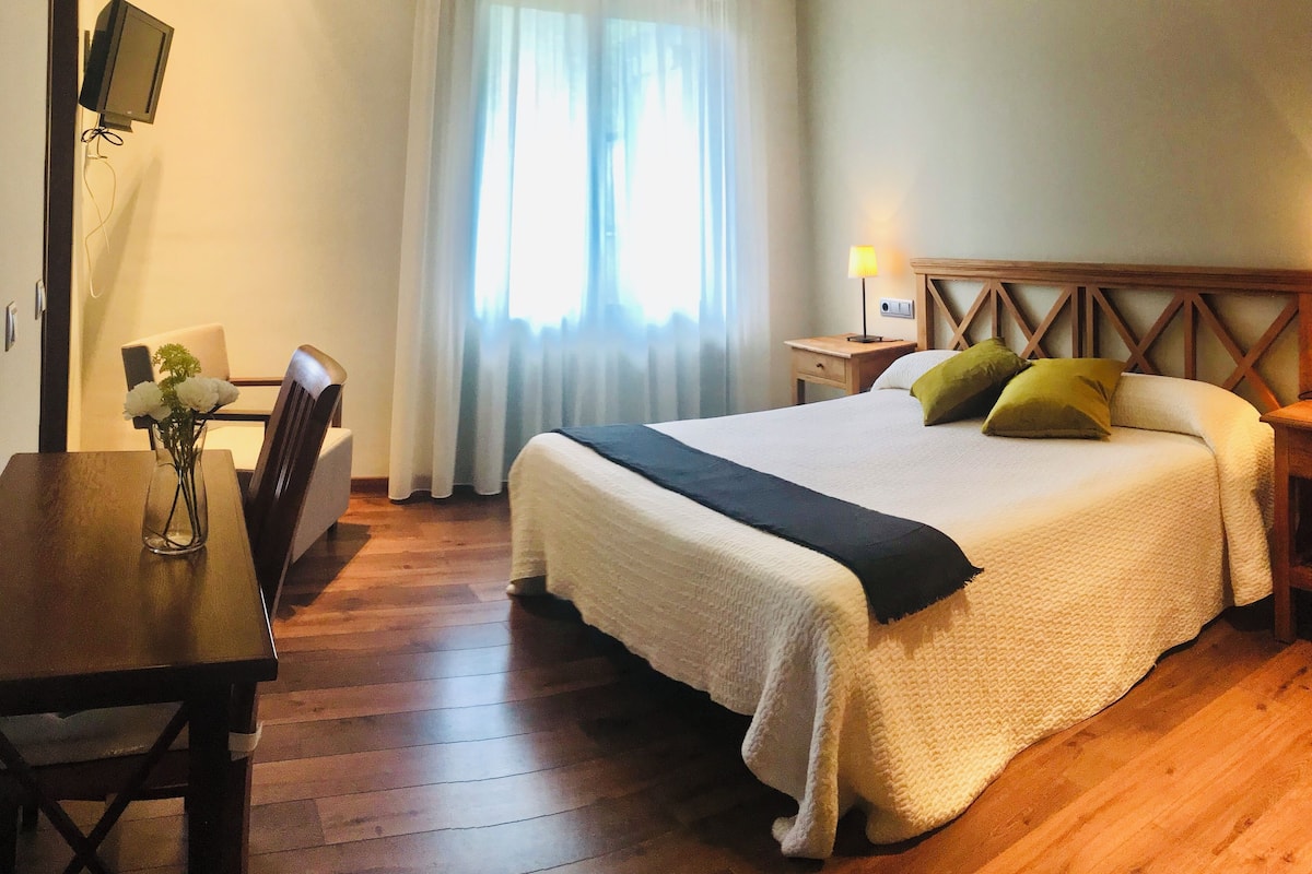 Una habitación de hotel con una cama grande, un escritorio de madera, una silla, un televisor montado en la pared y una ventana con cortinas.