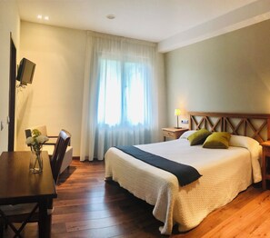 Una habitación de hotel con una cama grande, un escritorio de madera, una silla, un televisor montado en la pared y una ventana con cortinas.