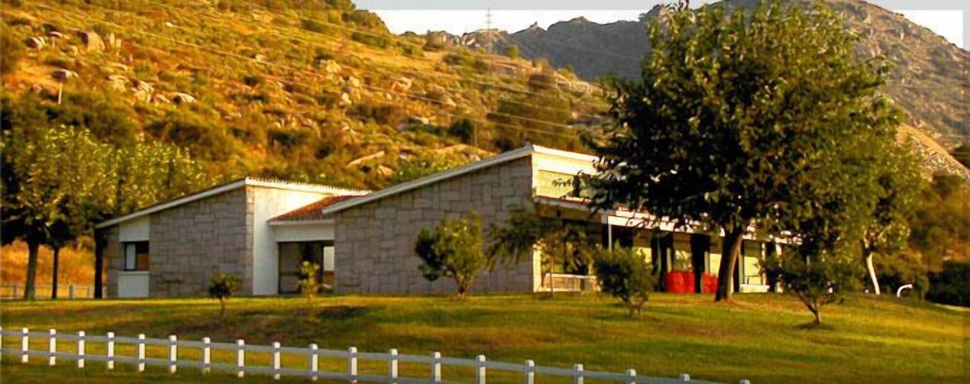 Un edificio de piedra de dos pisos con techo plano, rodeado por una cerca de piquetes blancos, con un paisaje montañoso de fondo.