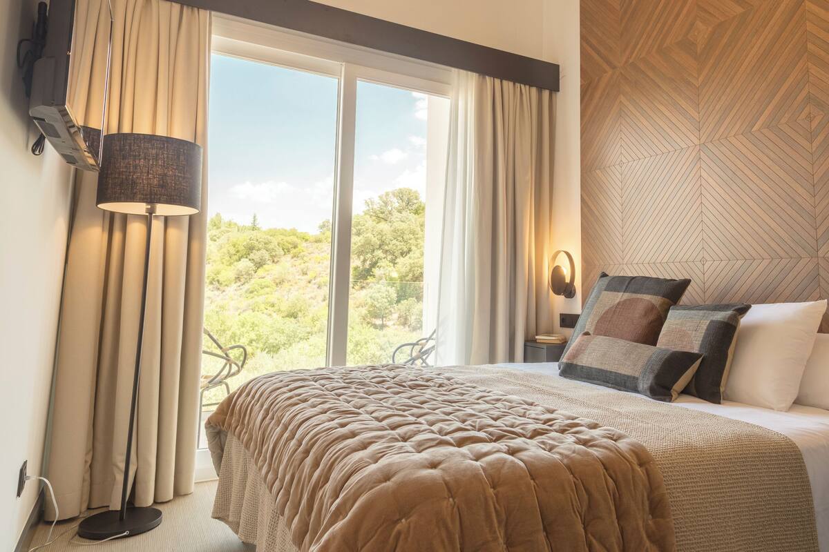 A bedroom with a large bed, a flat-screen TV mounted on the wall, a floor lamp, and a view of the outdoors through a large window.