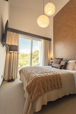 Ein Schlafzimmer mit einem großen Bett, einem flachbildfernseher an der Wand, einer Stehlampe und Blick ins Freie durch ein großes Fenster.