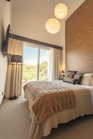 Un dormitorio con una cama grande, un televisor de pantalla plana montado en la pared, una lámpara de pie y vistas al exterior a través de un ventanal amplio.