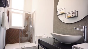 Et moderne badeværelse med et glasbadeværelsesafskærmning, et hvidt toilet, en stor hvid vask og et rundt spejl med hylder.