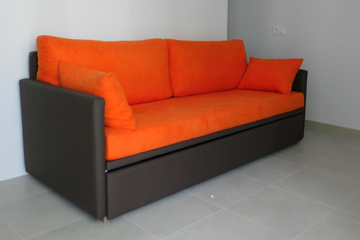 A dark brown sofa with orange cushions in a room with white walls and tiled flooring.