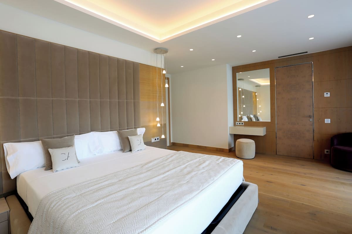 A modern bedroom with a large bed, wooden headboard, bedside lamps, a mirror, and a wooden floor.