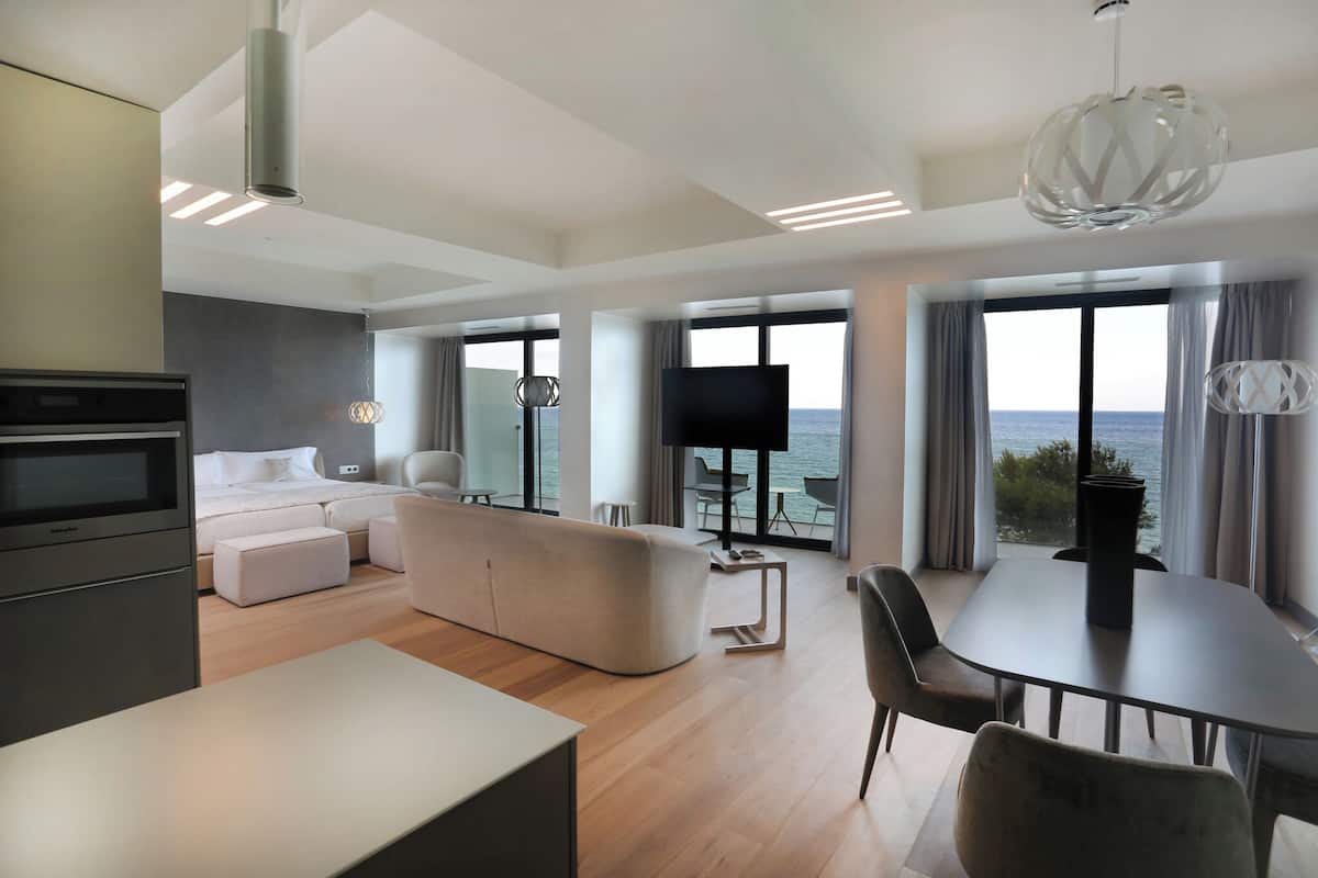 A modern hotel room with a large bed, a sofa, a dining table, and a TV. The room has large windows with a view of the sea.