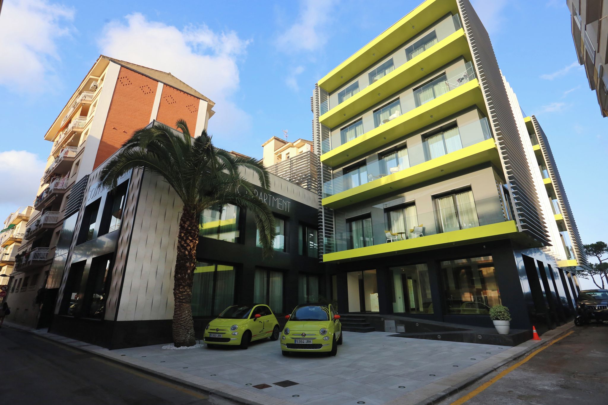 Un edifici modern amb façana de vidre, dos cotxes grocs aparcats al davant i una palmera propera.