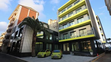 Een modern gebouw met een glazen gevel, twee gele auto's ervoor geparkeerd, en een palmboom in de buurt.