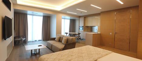 A modern hotel room with a large bed, a sofa, a desk, and a kitchenette.