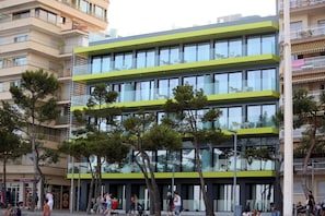 A modern multi-story building with balconies and large windows, surrounded by trees and people. - Cosmo Apartments Platja D'aro (Castell-Platja d'Aro)