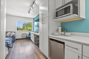 A modern kitchen with white cabinets, a dishwasher, and a microwave. There is a window providing a view of greenery outside. - 
Chic 1-bedroom Micro Studio Apartment (Cedar Falls)