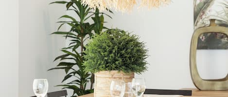 Een eetgedeelte met een geweven hanglamp, een potplant en een tafel gedekt voor twee personen, met wijnglazen en borden.