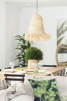 Zona pranzo con lampada a sospensione intrecciata, una pianta in vaso e tavolo apparecchiato per due con bicchieri da vino e piatti.