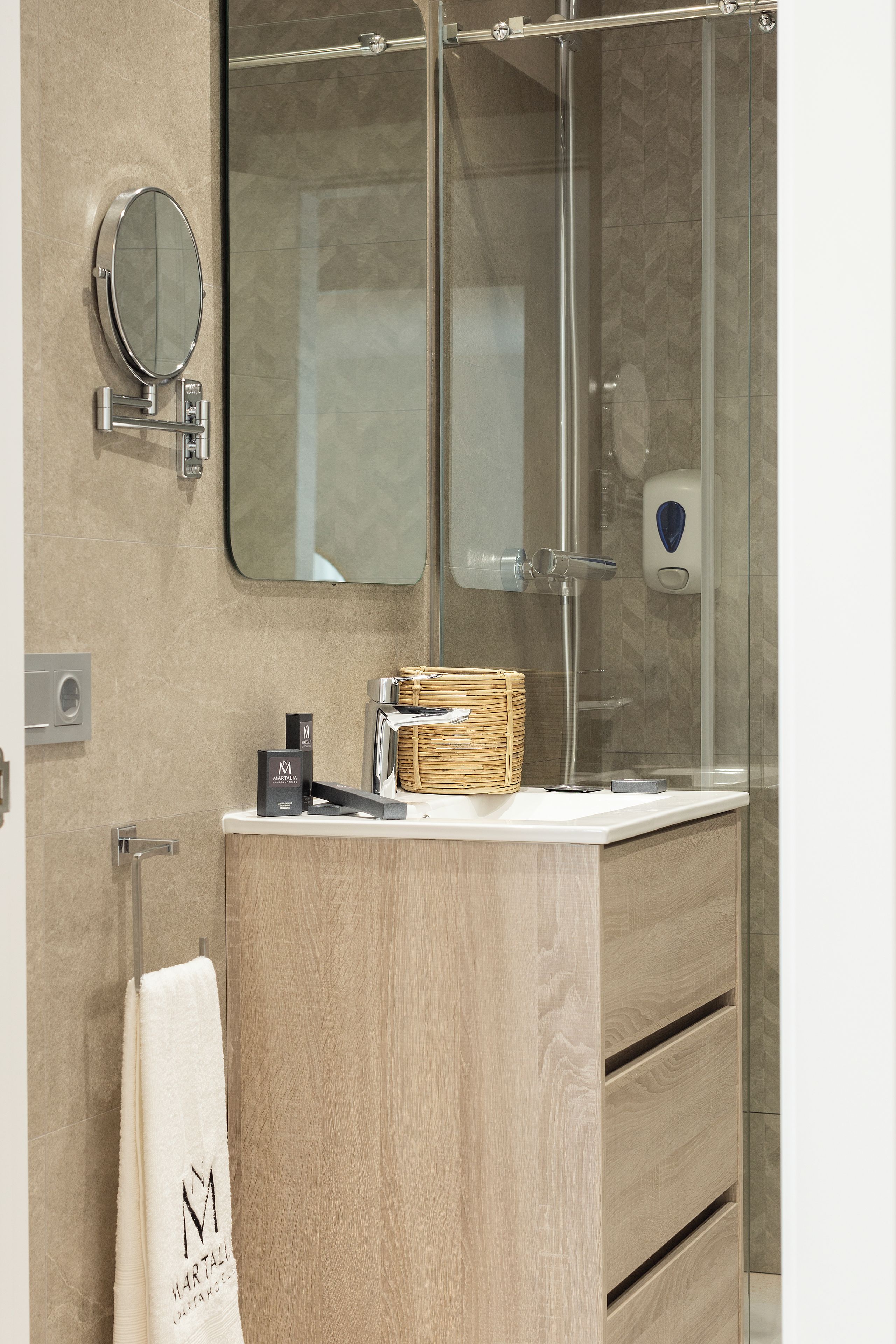 A modern bathroom with a wooden vanity, a glass shower enclosure, a round mirror, and a towel rack.