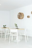 A modern dining area with white chairs and a round table, featuring woven wall art and potted plants.